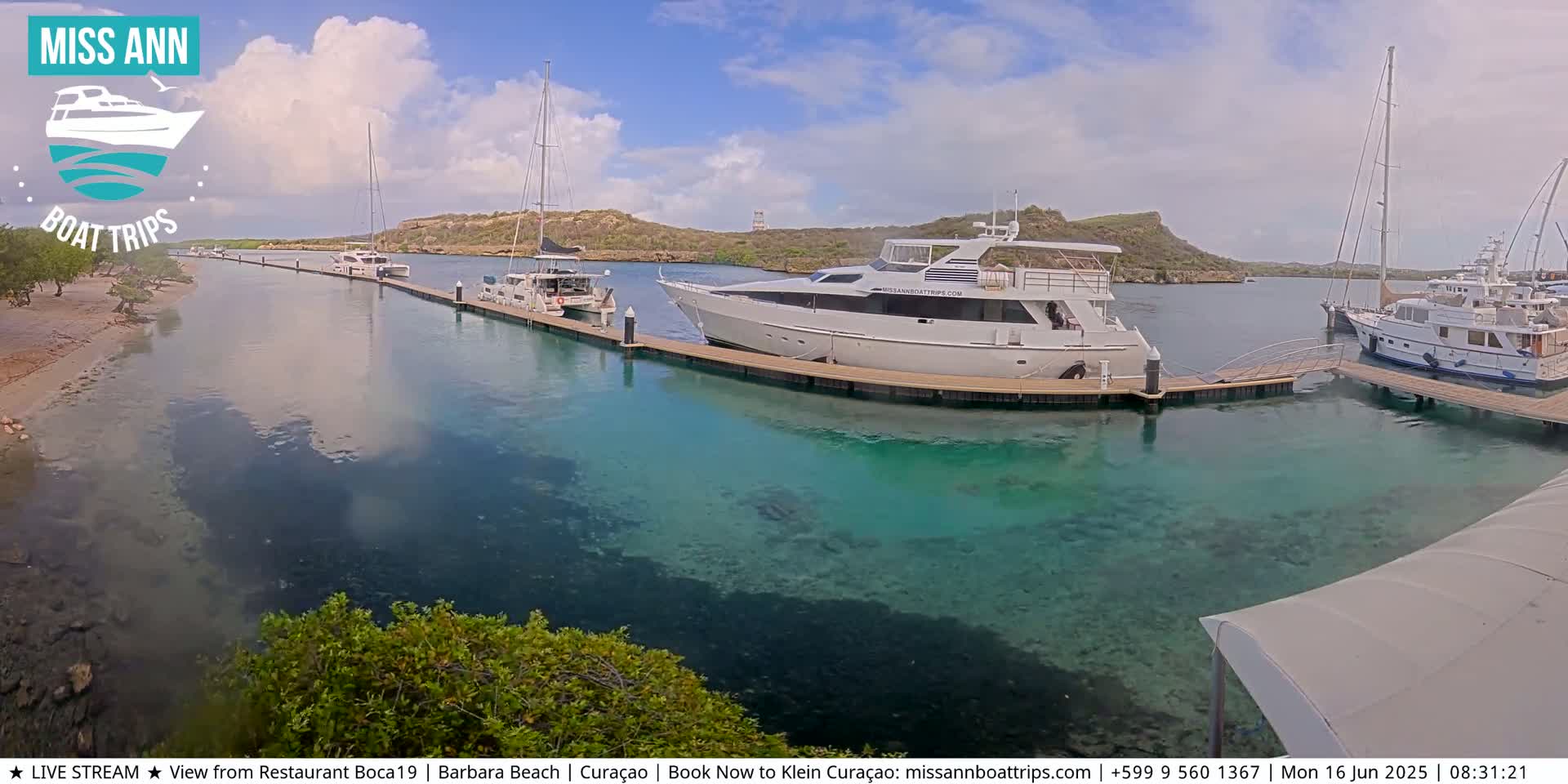 Several yachts and sailboats are docked in a calm, clear marina under a partly cloudy sky.