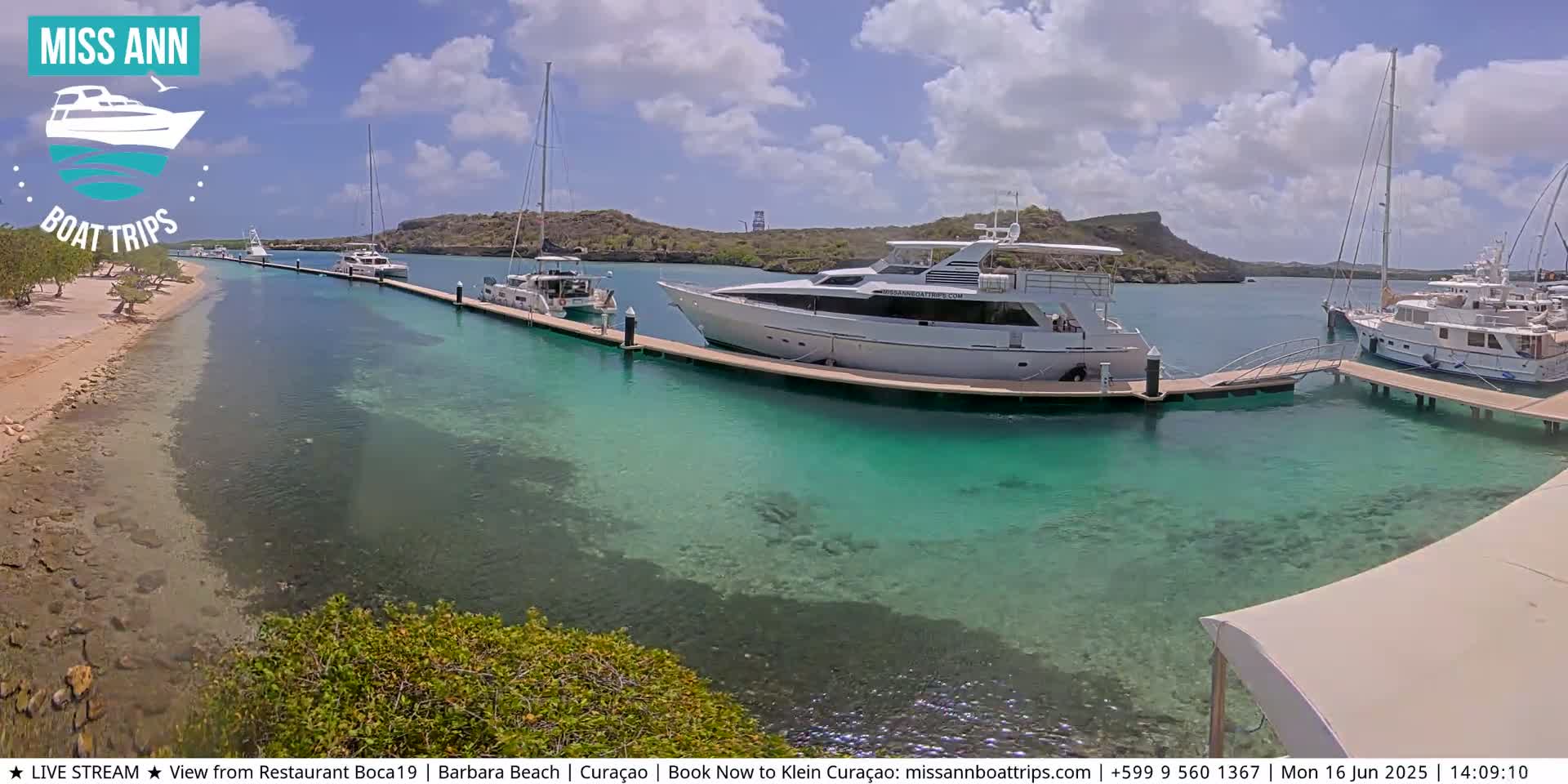 Several boats are docked at a marina with clear, shallow turquoise water, a sandy beach, and low-lying hills in the background under a partly cloudy sky.