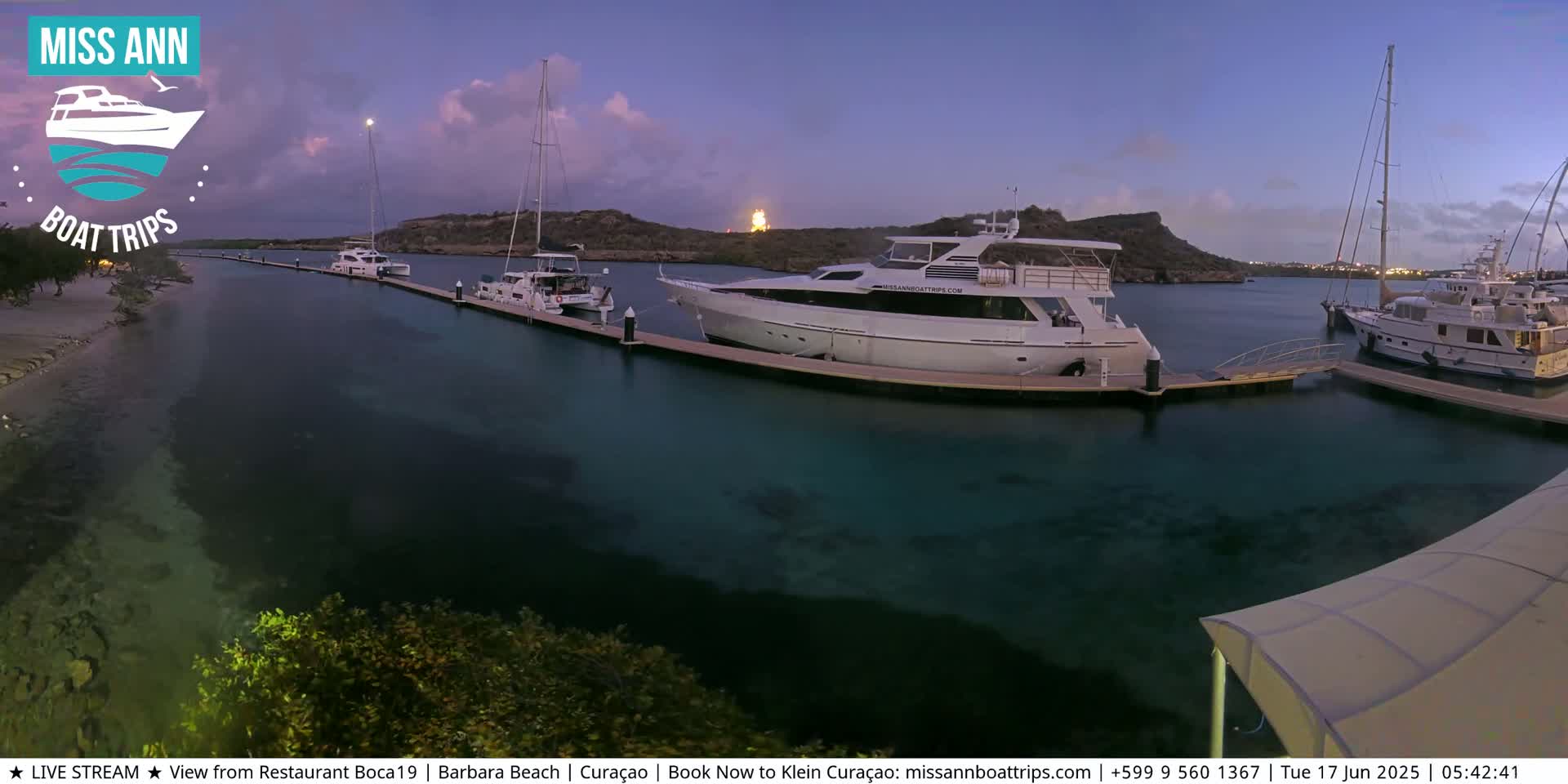 Several yachts and sailboats are docked at a marina at twilight, with clear, shallow turquoise water visible.