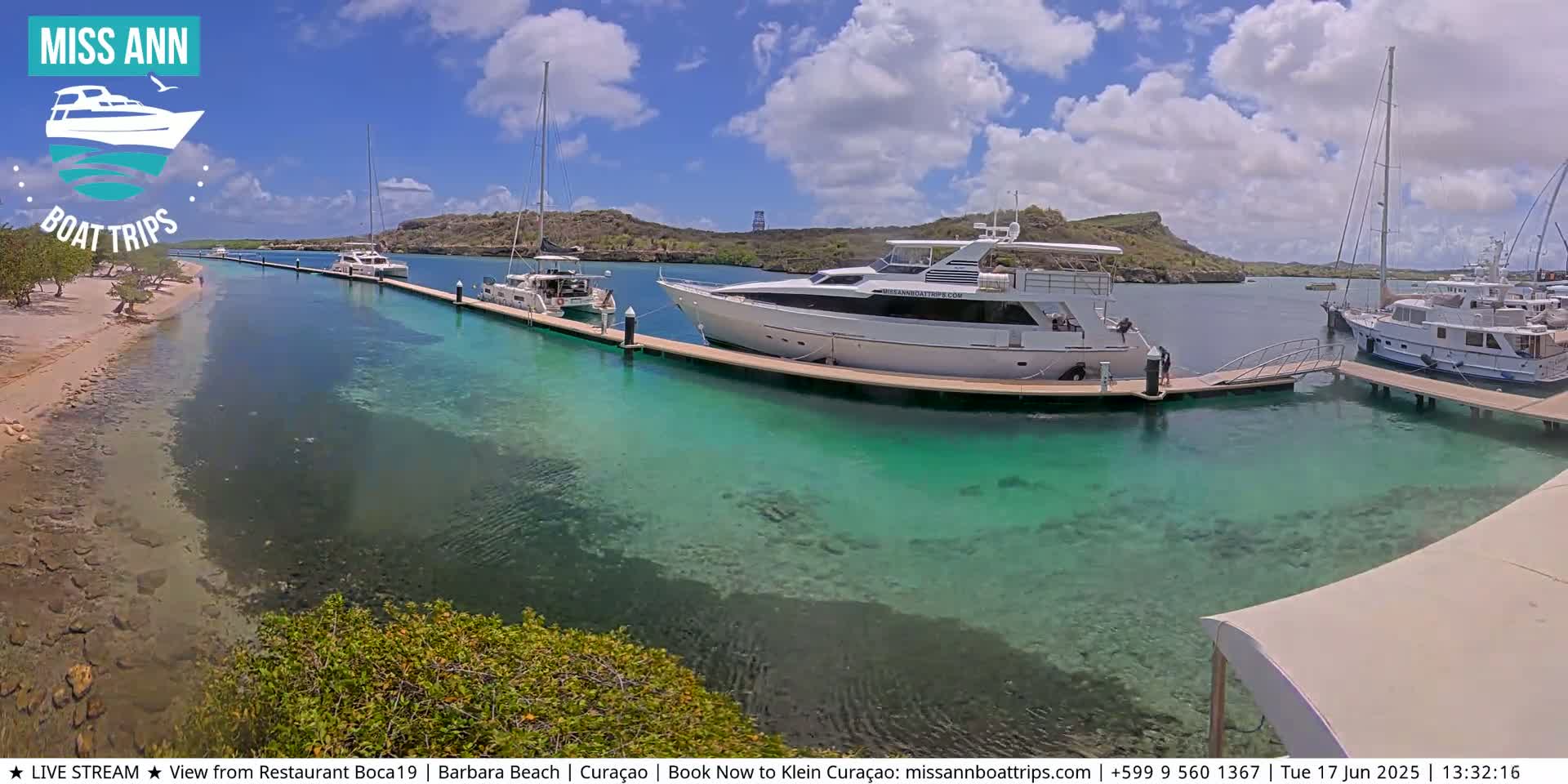 Several yachts and sailboats are docked at a marina with clear, shallow turquoise water, under a partly cloudy sky.