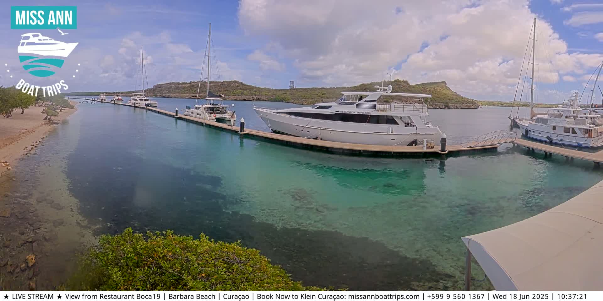 Several yachts and sailboats are moored at a marina on a sunny day with partly cloudy skies and clear, shallow turquoise water.