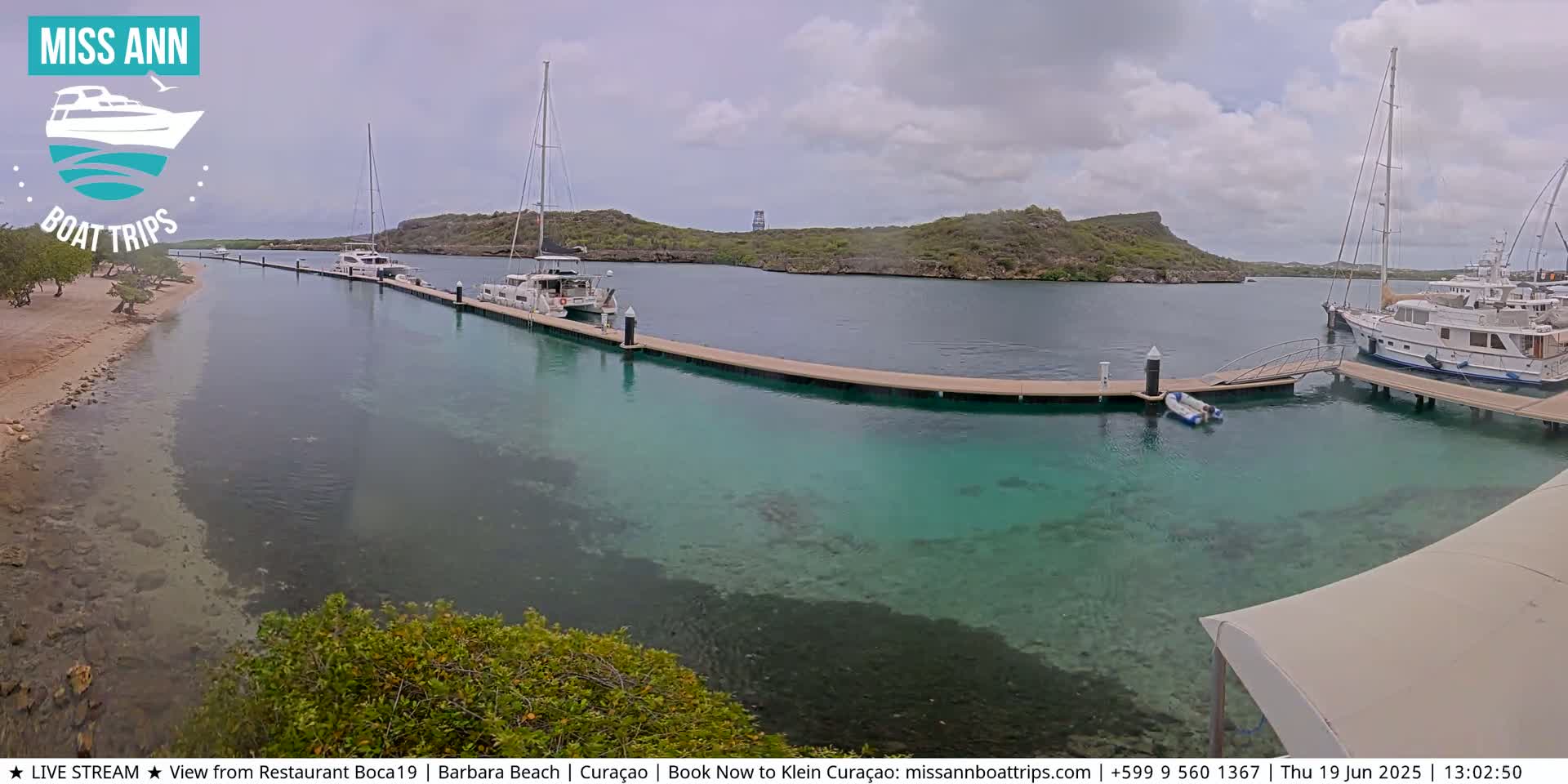 Several boats are docked at a marina with clear, shallow turquoise water, under a partly cloudy sky.