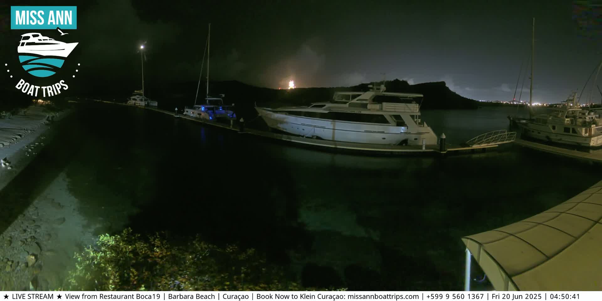 Several yachts are moored at a marina at night, under a partly cloudy sky with a distant light source visible in the background.