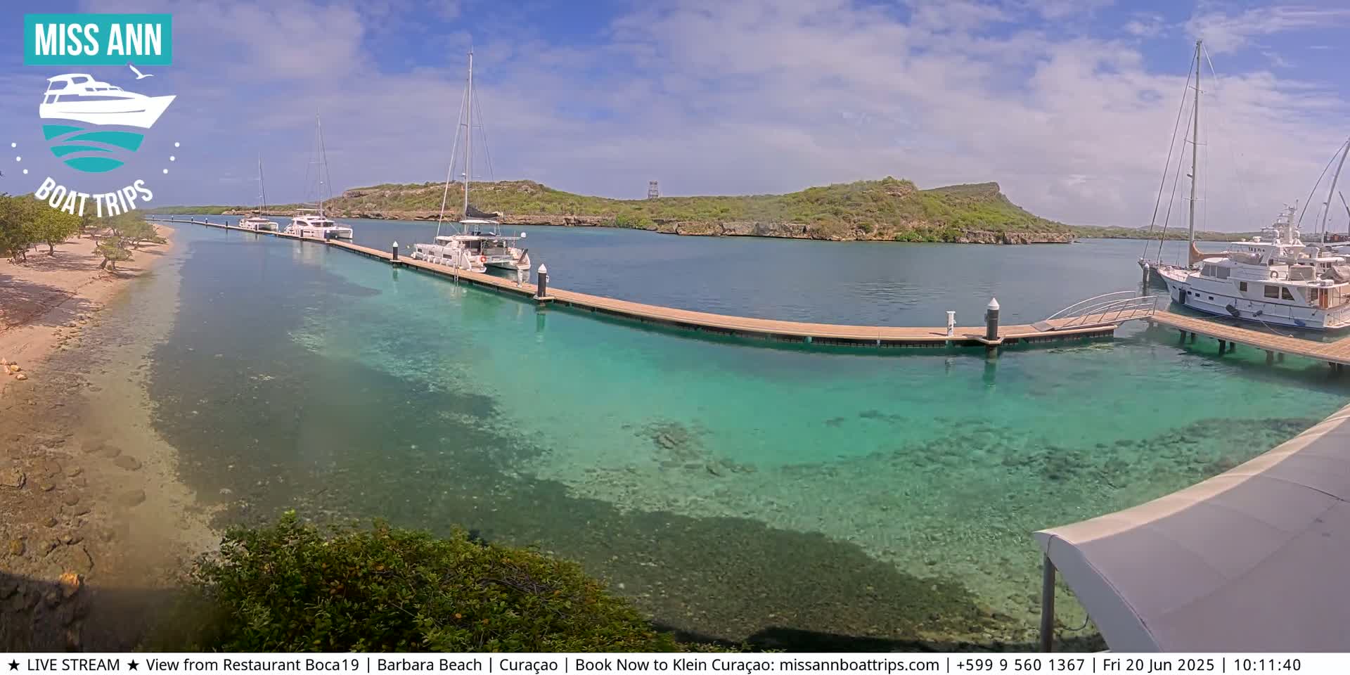 Several boats are docked at a marina with clear, shallow turquoise water, a sandy beach, and lush green hills in the background under a sunny sky.