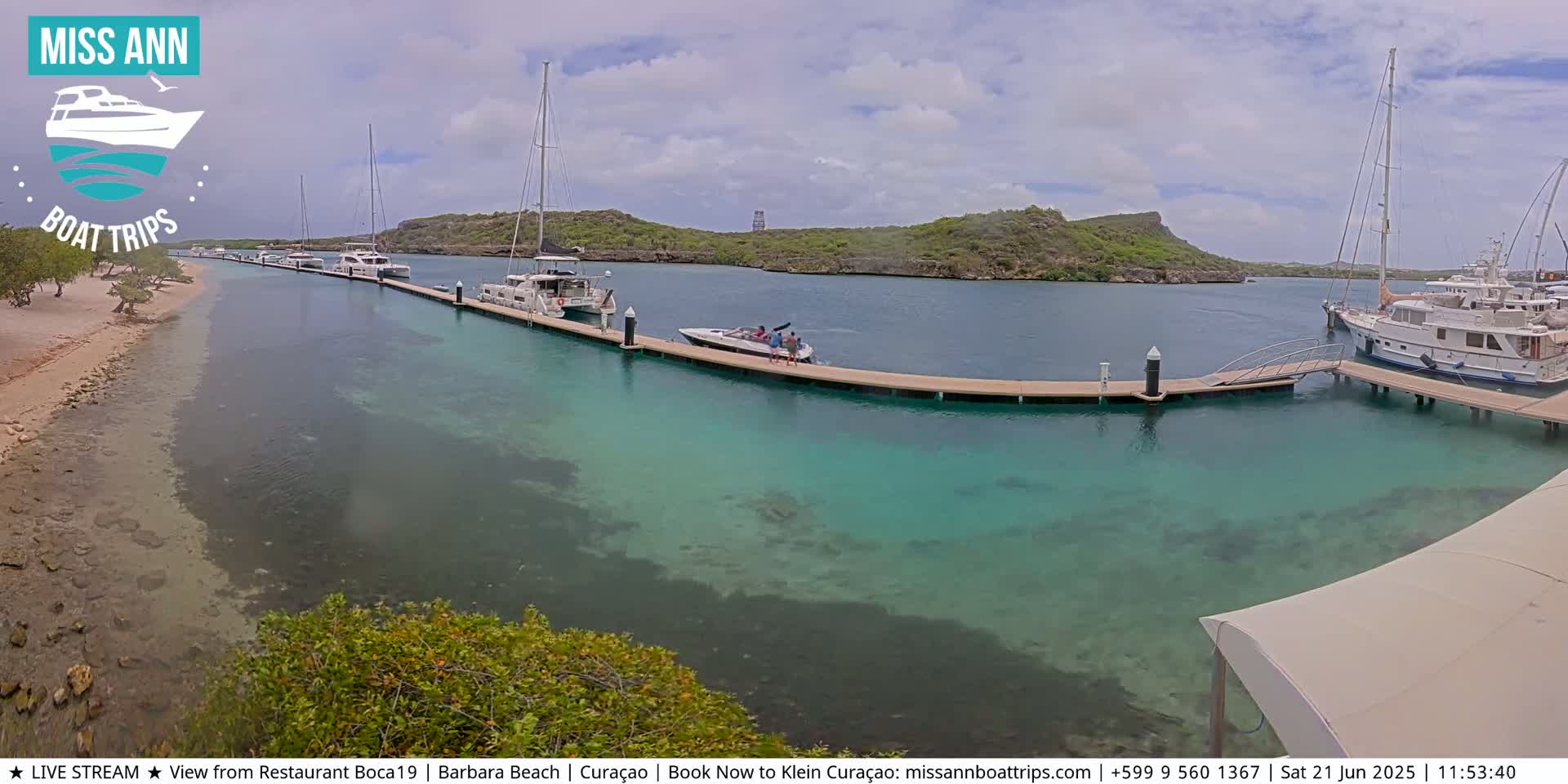Several boats are docked at a marina with clear, shallow turquoise water, under a partly cloudy sky.