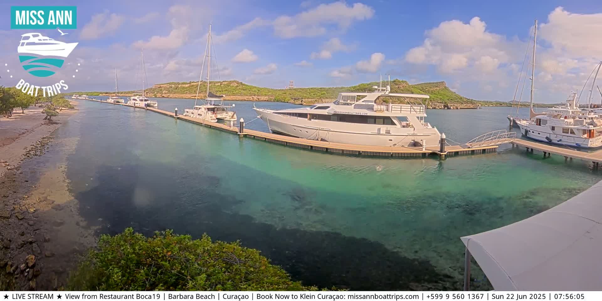 Several yachts and sailboats are docked in a marina on a sunny day with clear, shallow turquoise water.