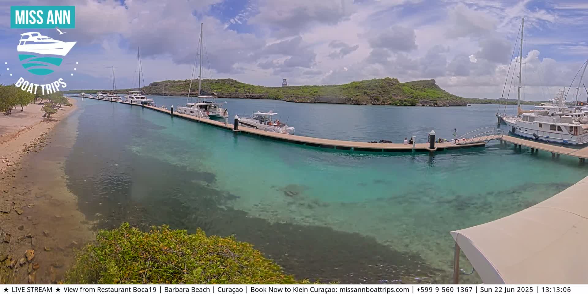 Several boats are docked at a marina with clear, shallow turquoise water, under a partly cloudy sky.