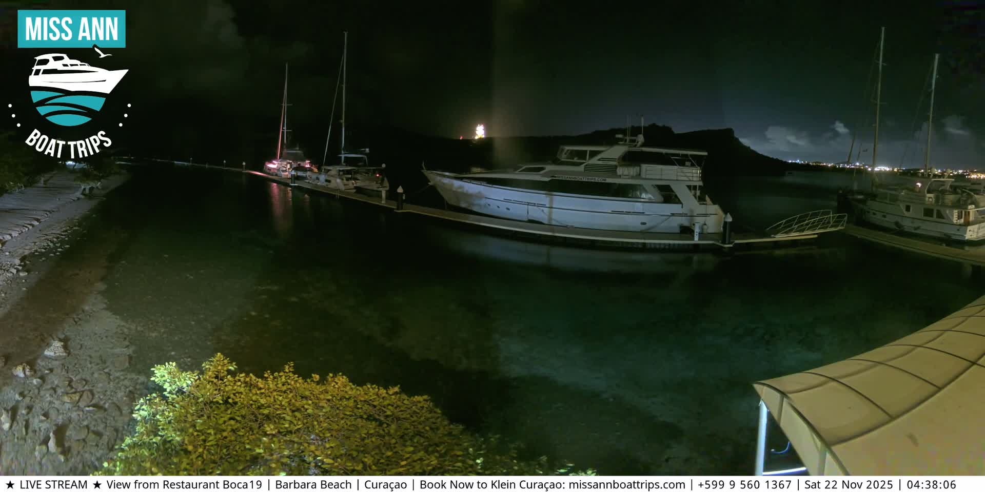 At night, several boats are docked along illuminated piers in a calm marina under a clear sky, with the water reflecting lights from the boats and a distant shore.