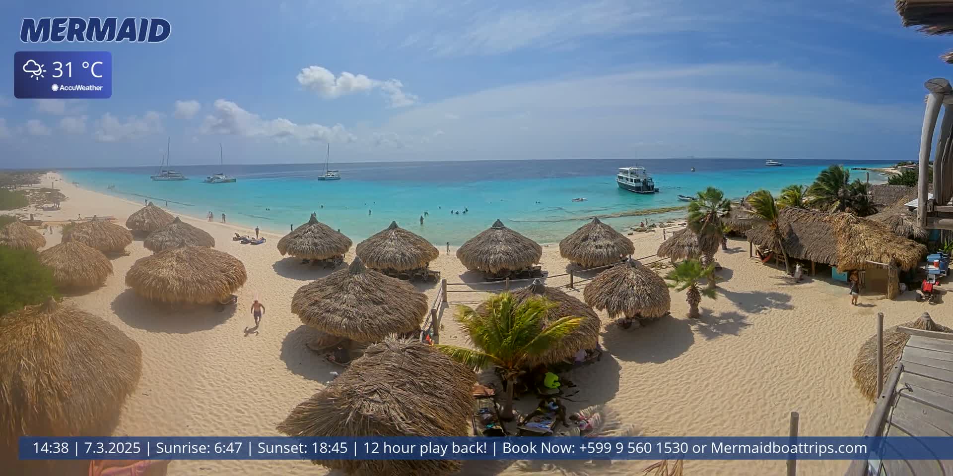 A sunny beach with turquoise water, numerous thatched-roof huts, and several boats offshore.