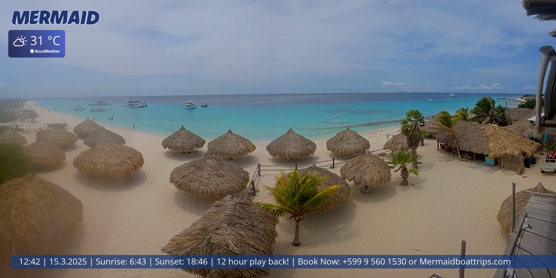 A beach with numerous thatched-roof umbrellas, boats in the turquoise water, and palm trees under a partly cloudy sky.