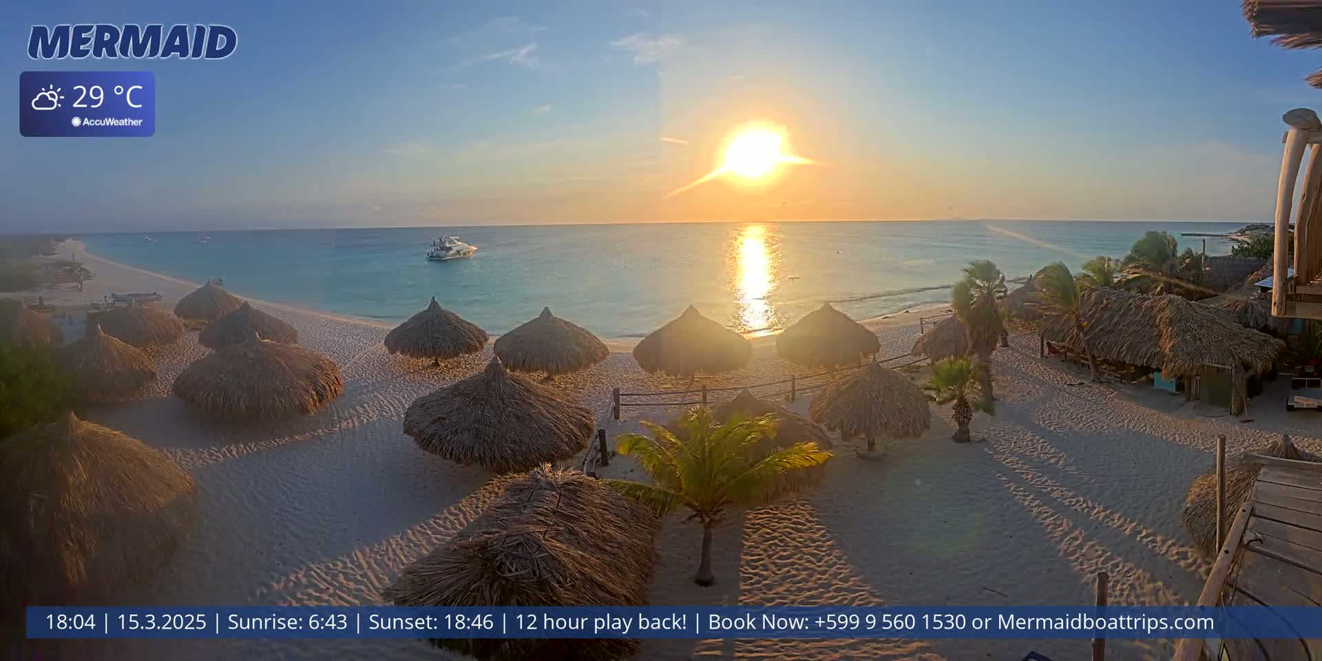A beach scene at sunset shows several thatched-roof huts on a sandy beach next to a calm, turquoise ocean.