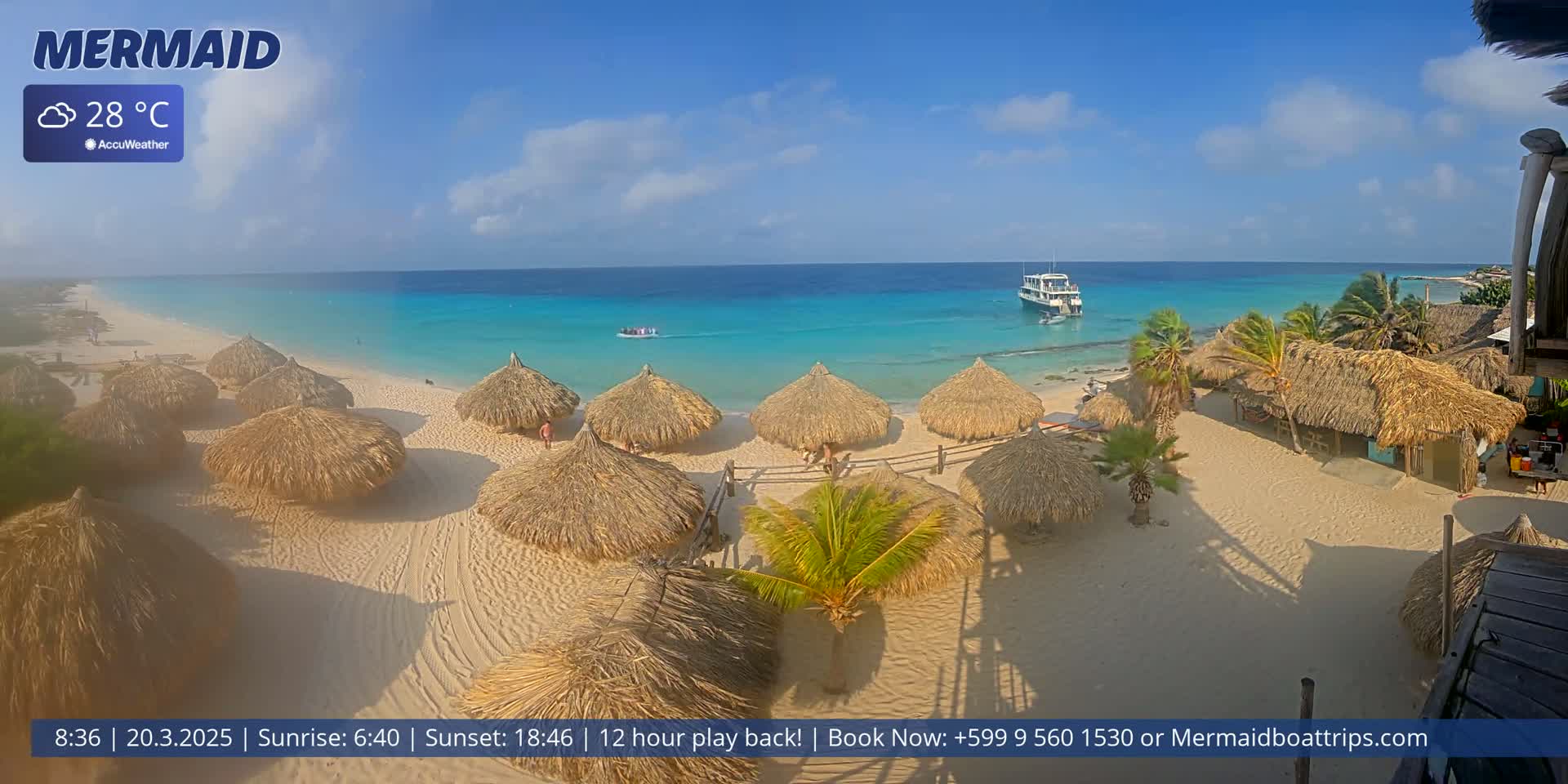 A sandy beach with numerous thatched-roof huts is pictured on a sunny day with clear turquoise water and a boat offshore.