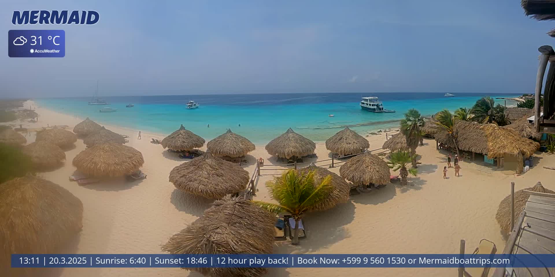 A beach with numerous thatched-roof huts, several boats offshore, and clear turquoise water under a sunny sky.