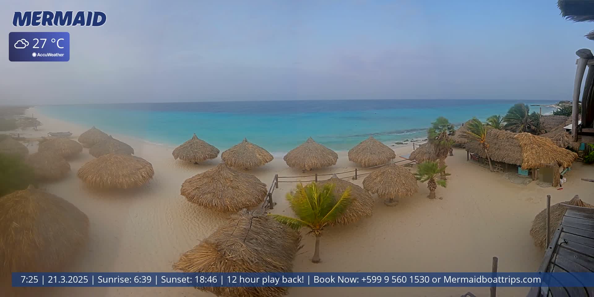 A hazy beach scene shows several thatched-roof huts on a white sand beach next to turquoise water under a cloudy sky.