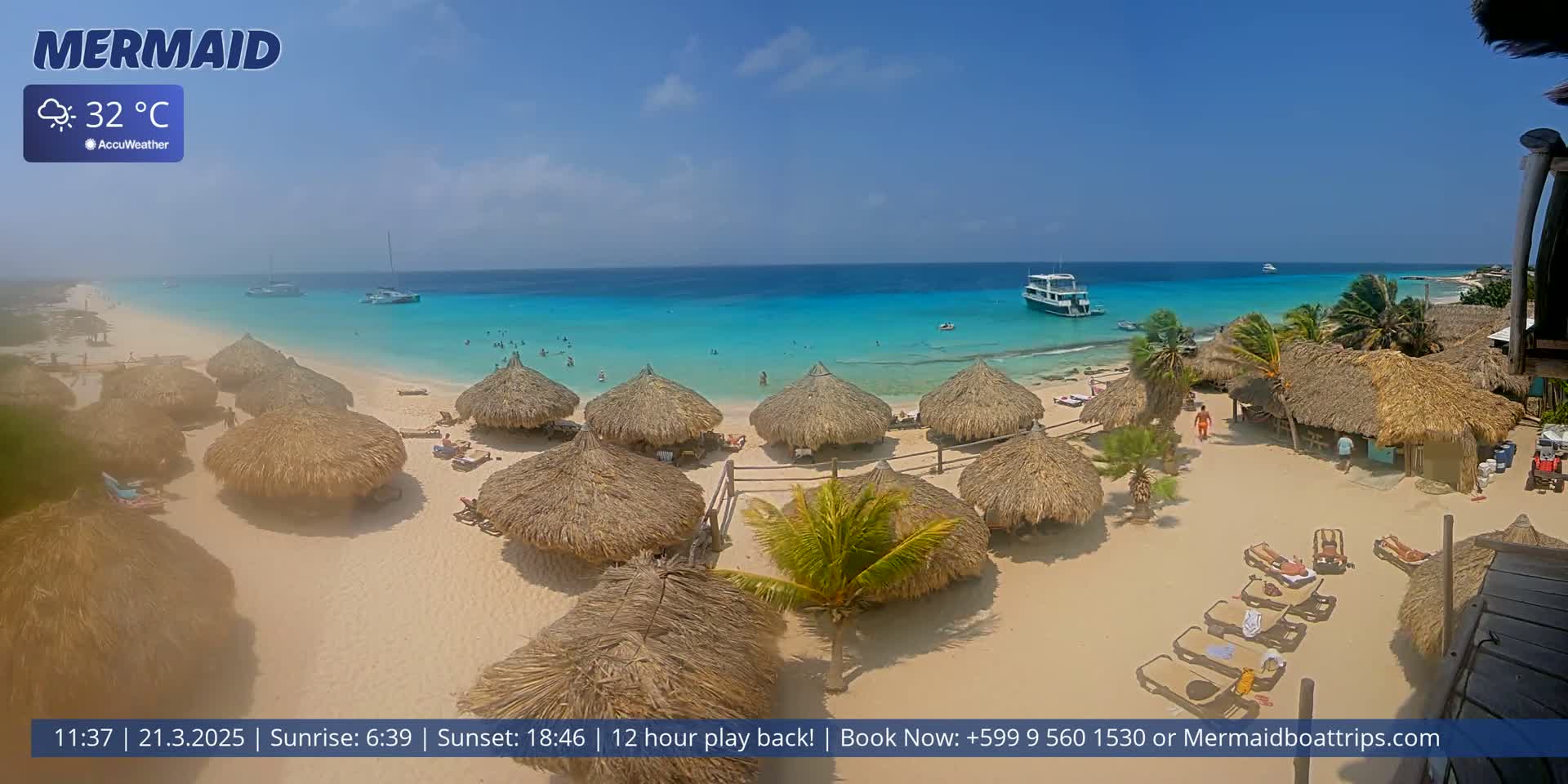 A sunny beach with turquoise water, numerous thatched-roof huts, and several people relaxing on lounge chairs and in the water.