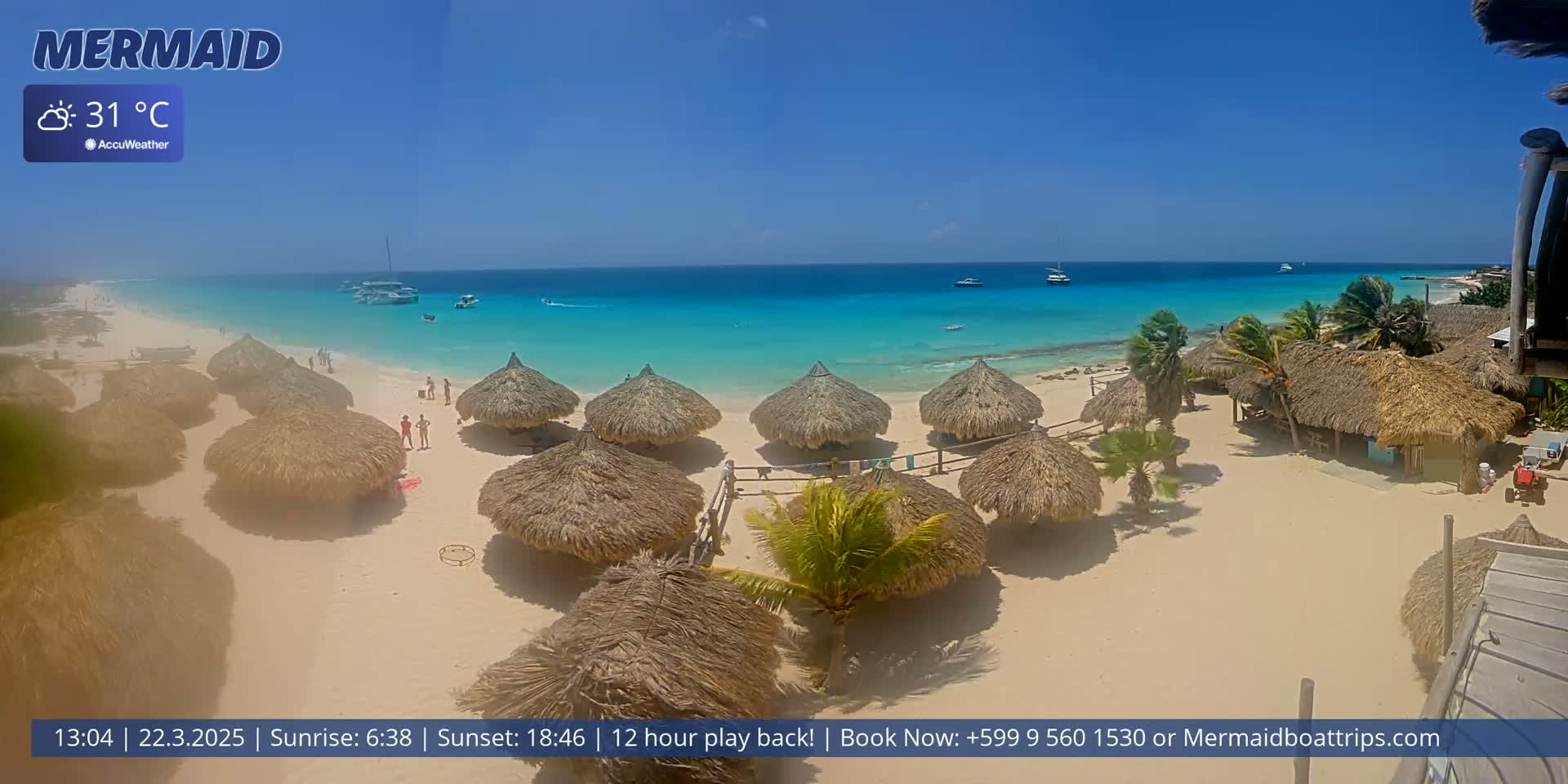 A sunny beach scene with numerous thatched-roof umbrellas, a few people, and several boats visible in the turquoise water under a clear blue sky.
