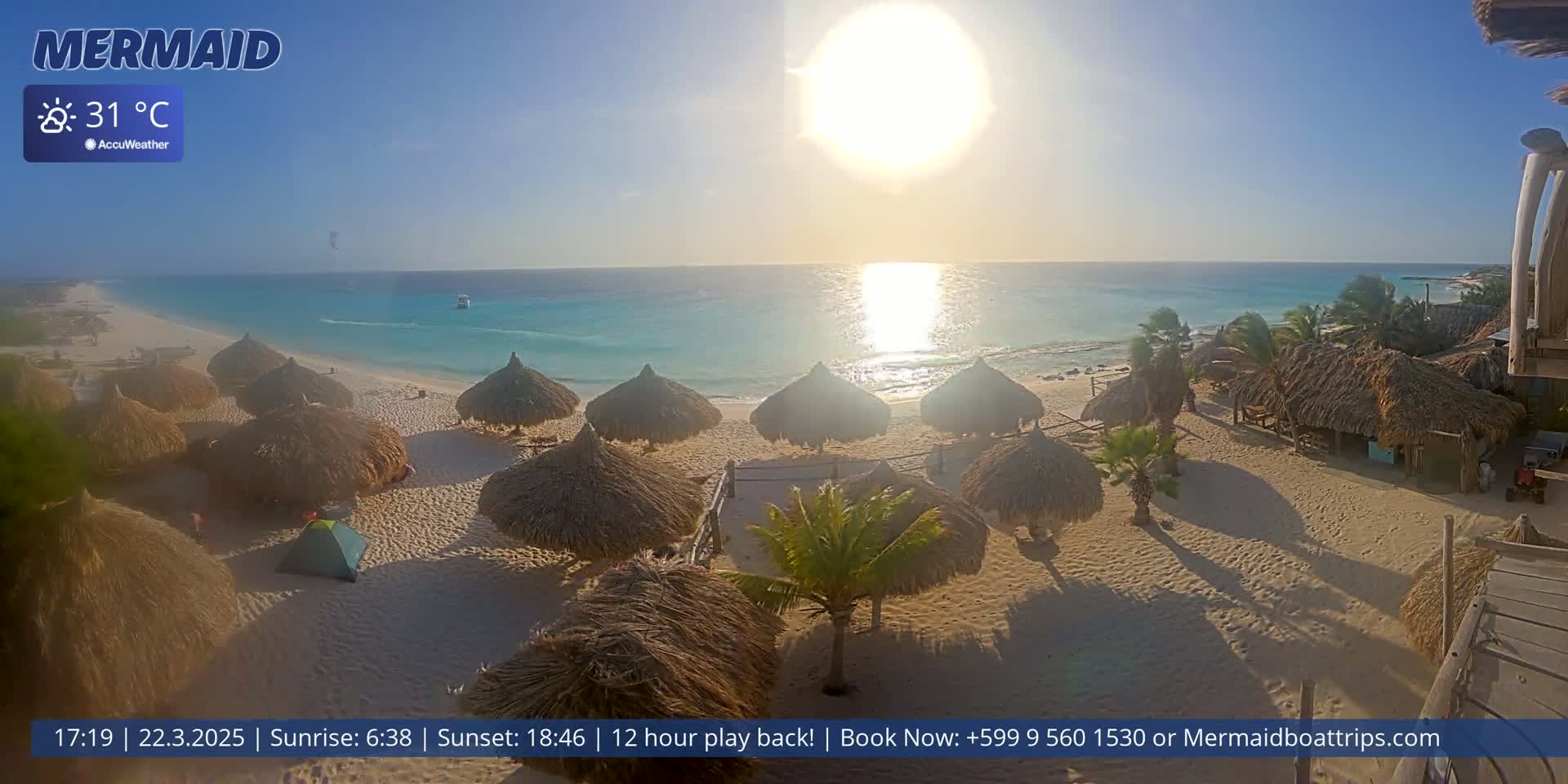 A sunny beach scene shows numerous thatched-roof beach umbrellas on a sandy shore, with calm turquoise water and a bright sun in the sky.