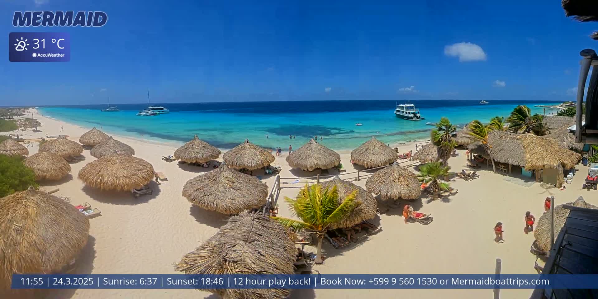 A beach with numerous thatched umbrellas, some people, and two boats in a clear turquoise ocean under a sunny sky.