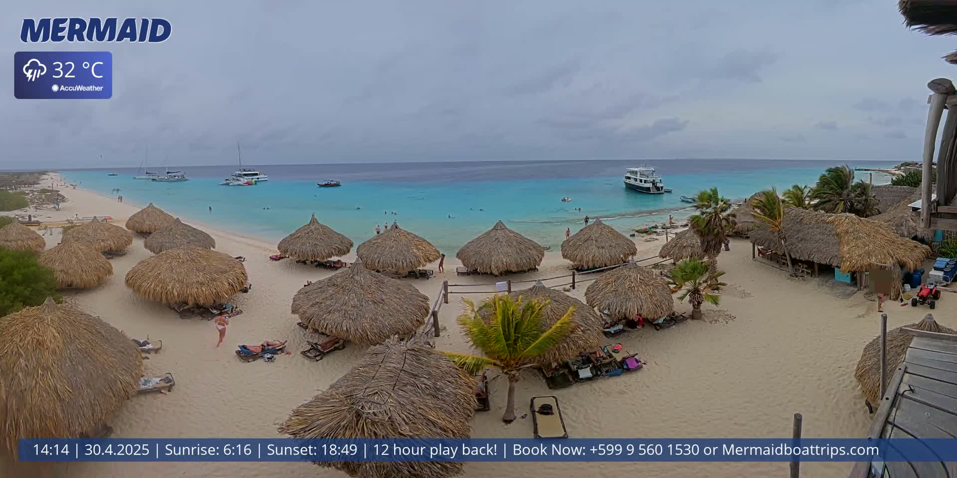 A sandy beach with numerous thatched-roof huts, a few people, and boats offshore on a partly cloudy day.