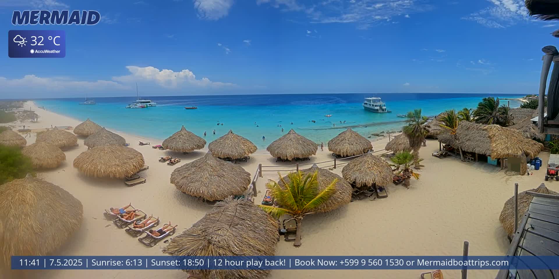 A beach with numerous thatched-roof huts, people relaxing on lounge chairs, and several boats offshore in clear, turquoise water under a partly sunny sky; the temperature is 32°C.