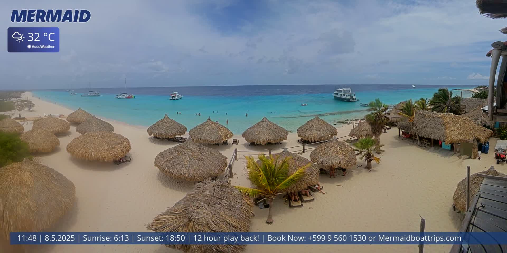 A sandy beach with numerous thatched-roof huts is shown under partly cloudy skies, with calm, turquoise water and several boats visible offshore.