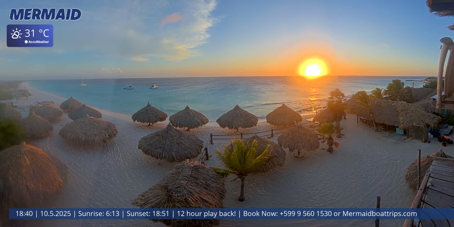 A beach scene at sunset shows several thatched-roof umbrellas on a sandy beach with calm turquoise water and a bright orange sun in the sky, with the air temperature at 31°C.