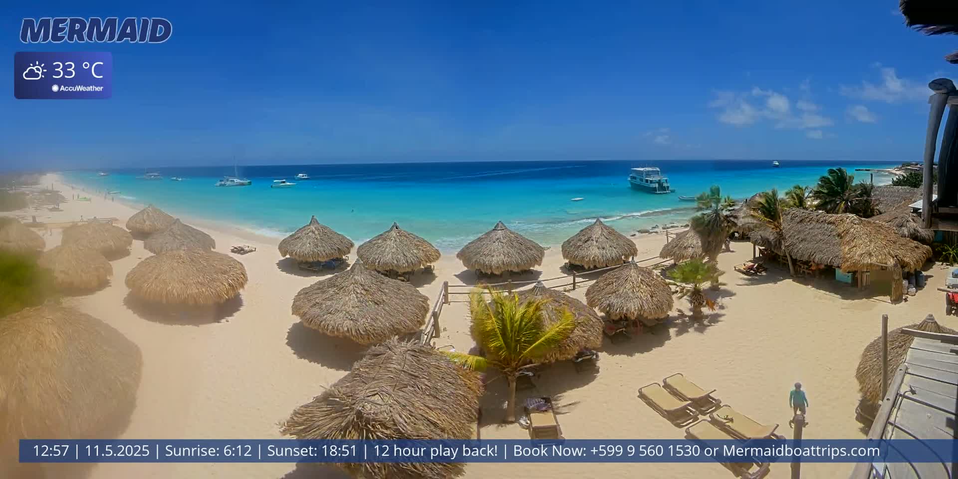 A beach with numerous thatched-roof umbrellas, a few boats offshore, and clear turquoise water under a bright sunny sky.