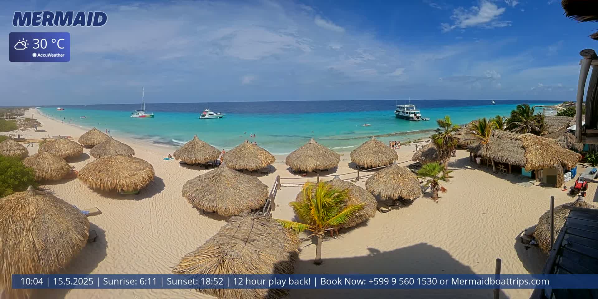 A sandy beach with numerous thatched-roof huts is situated next to clear turquoise water under a sunny sky; several boats are visible on the water.