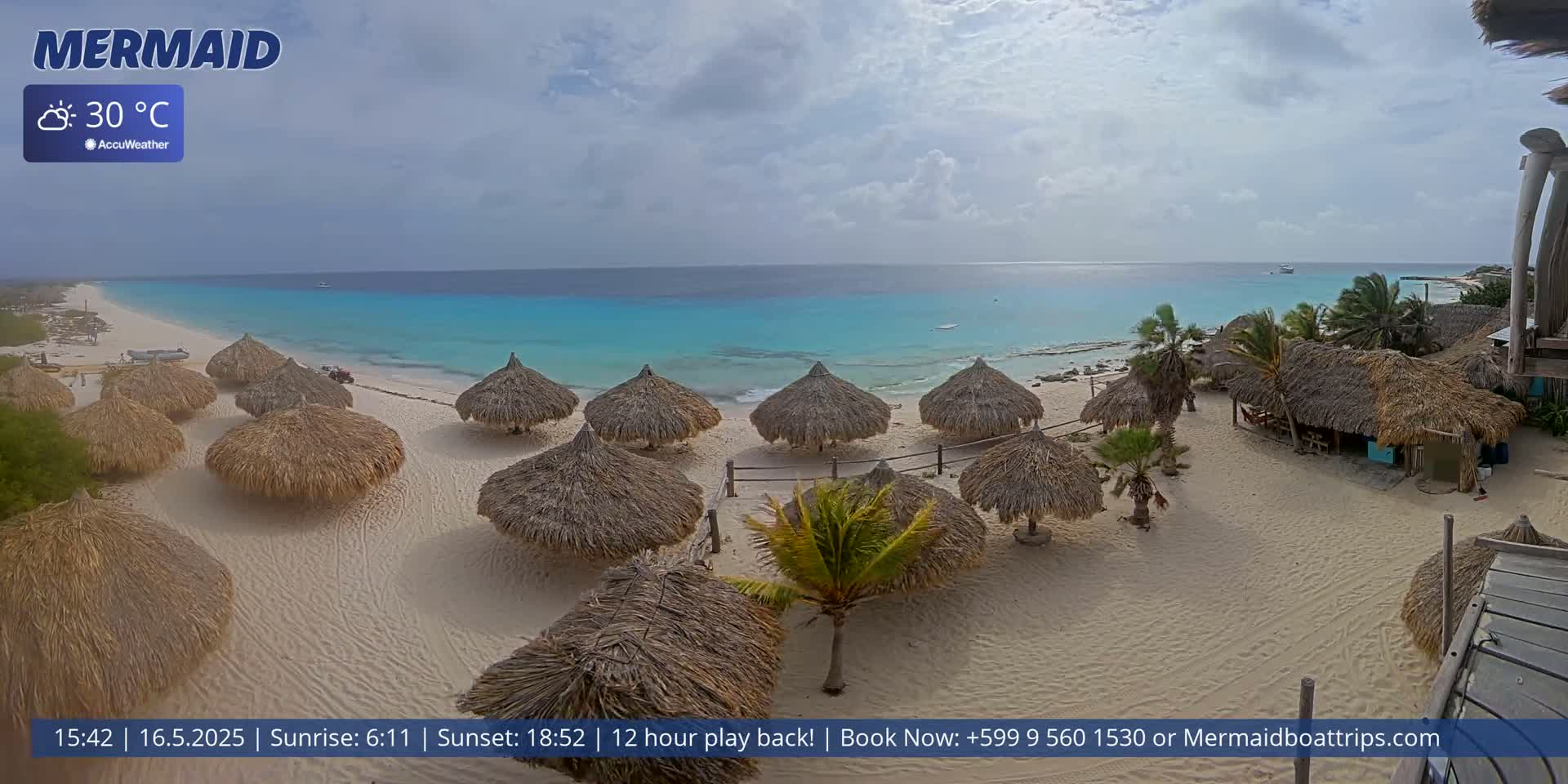 A partly cloudy day reveals a beach with numerous thatched umbrellas, turquoise water, and a few small buildings.