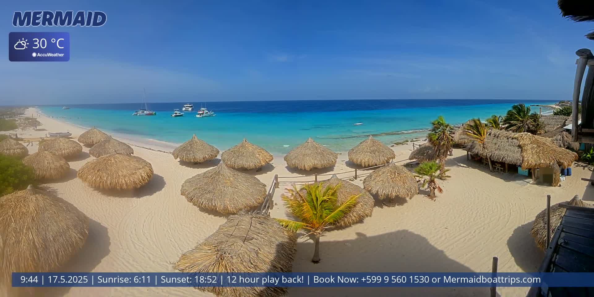 A beach with numerous thatched-roof huts, several boats offshore, and clear turquoise water under a sunny sky.