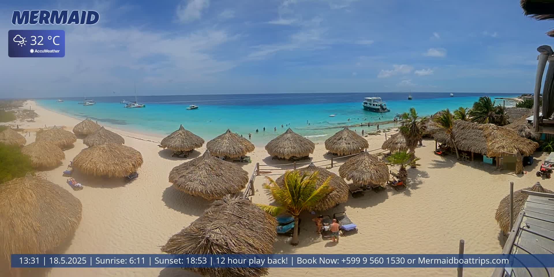 A sandy beach with numerous thatched-roof huts is shown under a sunny, clear sky; the turquoise ocean stretches beyond, with several boats and people swimming in the water.