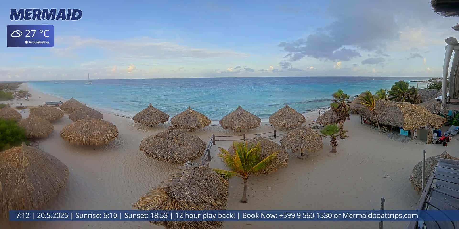 A sandy beach with numerous thatched-roof huts, palm trees, and a calm turquoise ocean under a partly cloudy sky.