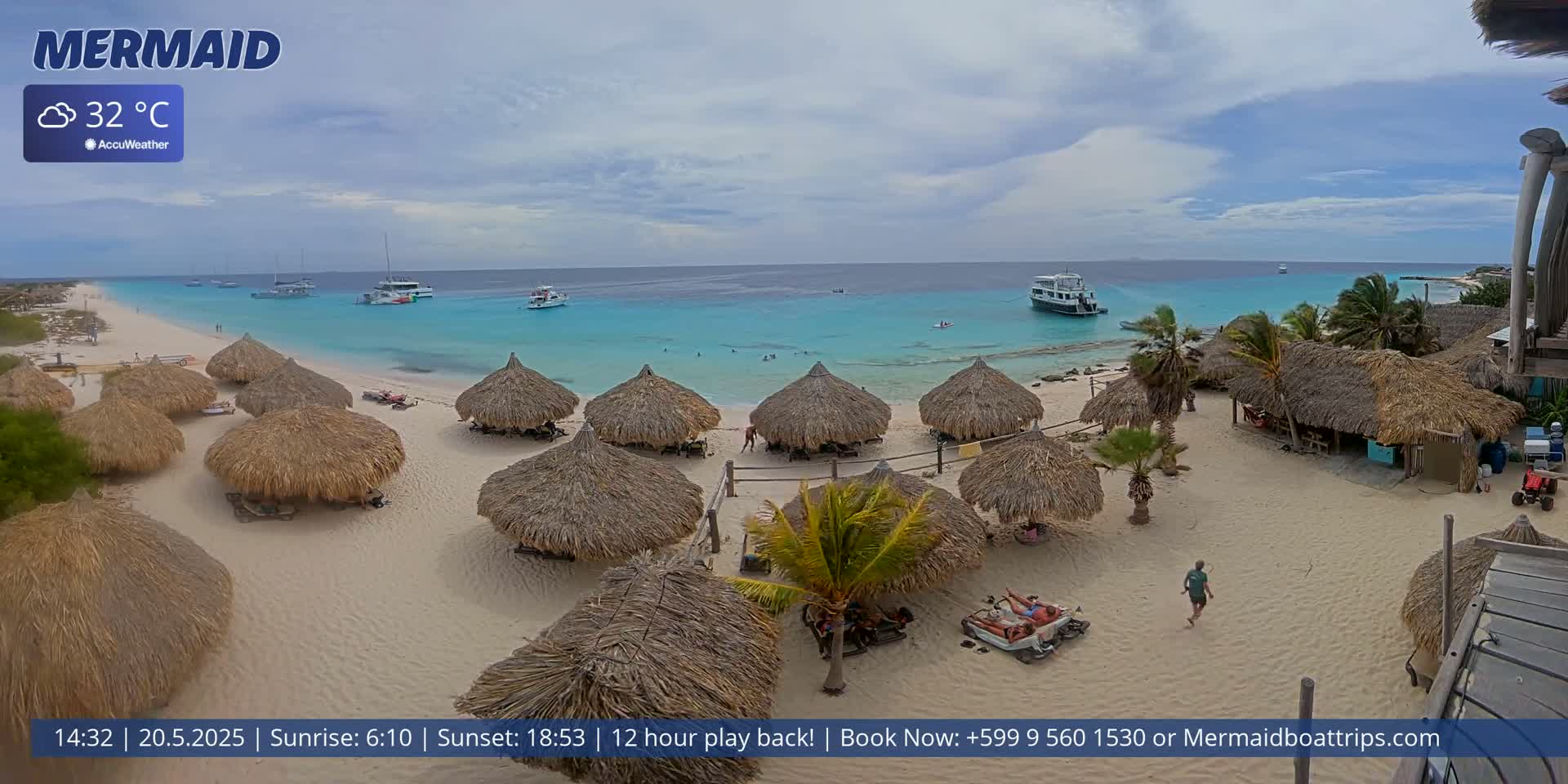 A beach with thatched-roof huts, several boats in the turquoise water, and partly cloudy skies.