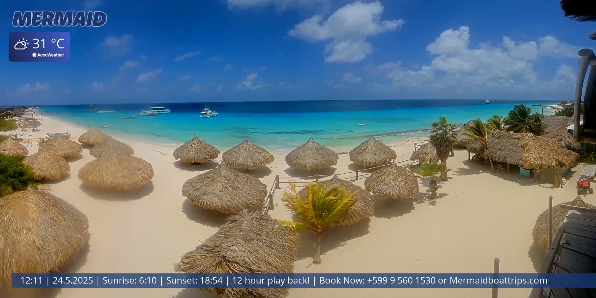 A beach with numerous thatched-roof huts, boats in the turquoise water, and a partly cloudy sky under sunny conditions.