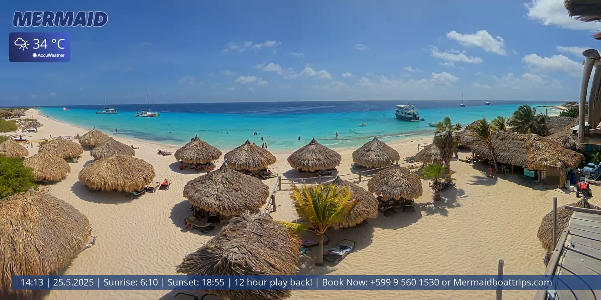 A beach with numerous thatched-roof huts, some people swimming and relaxing in the turquoise water, and boats visible offshore on a sunny day with a temperature of 34°C.