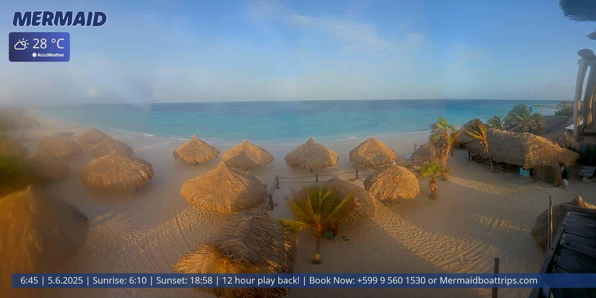 A sandy beach with numerous thatched-roof huts is situated next to a calm, turquoise ocean under a mostly clear sky; the temperature is 28°C.