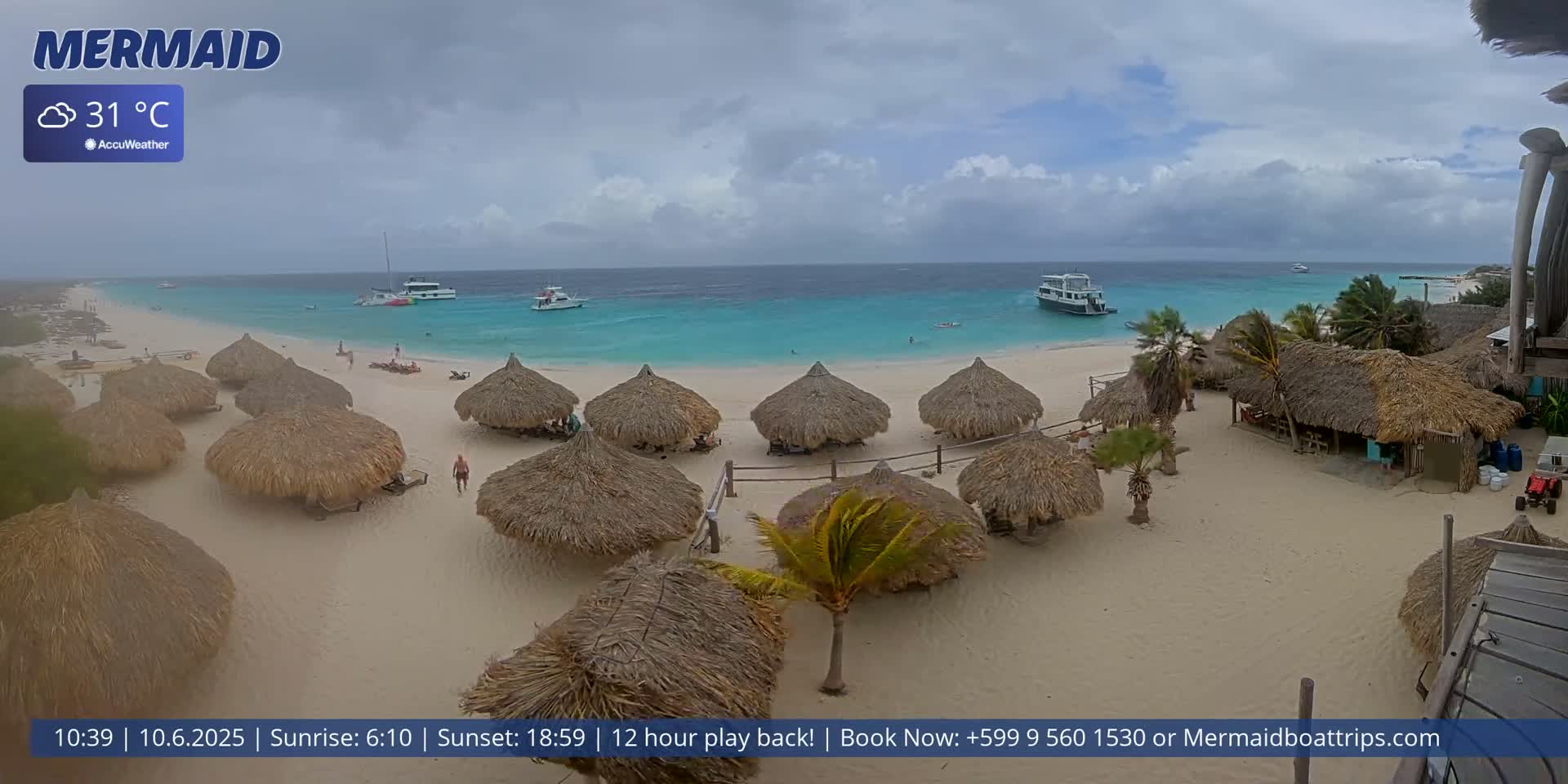 A sandy beach with numerous thatched-roof huts, several boats in the turquoise water, and cloudy skies.