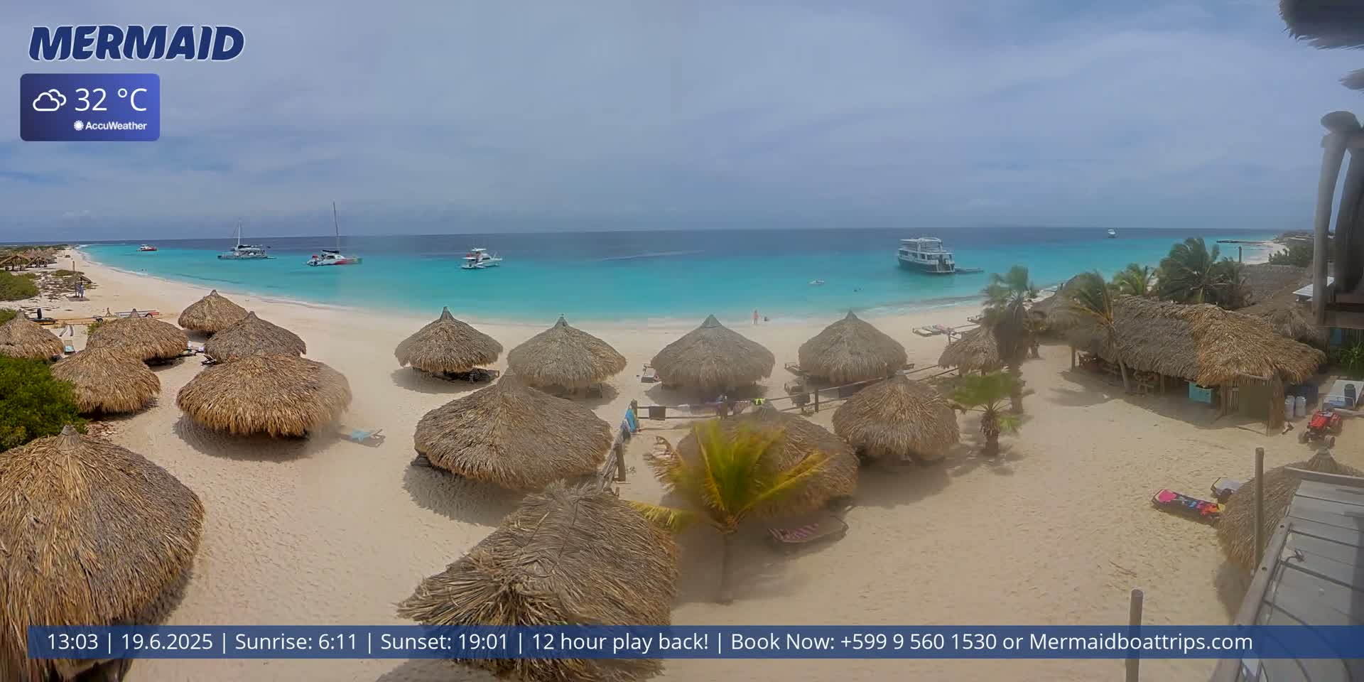 A beach with numerous thatched-roof huts, several boats in the turquoise water, and a partly cloudy sky.