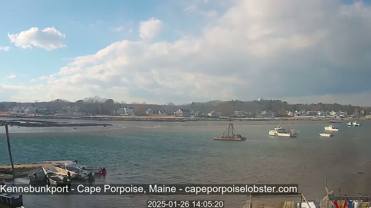 A partly cloudy day reveals a calm bay with several small boats near a shoreline of houses, some with snow on the ground.