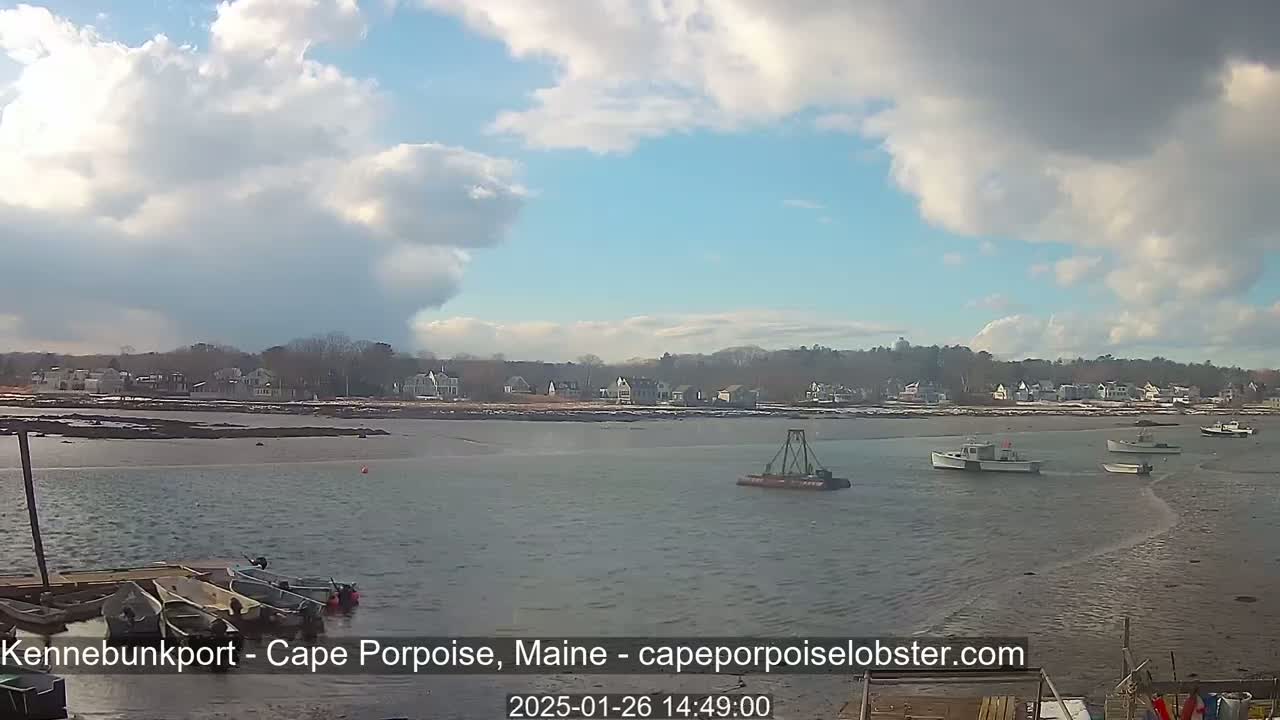 A partly cloudy day shows several small boats in calm water near a shoreline with houses and trees, some areas of which appear to have patches of snow.
