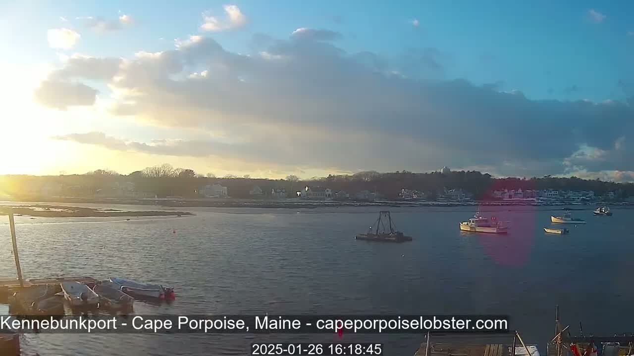 A calm body of water with several small boats is seen at sunset, with a shoreline of houses and trees in the background under a partly cloudy sky.