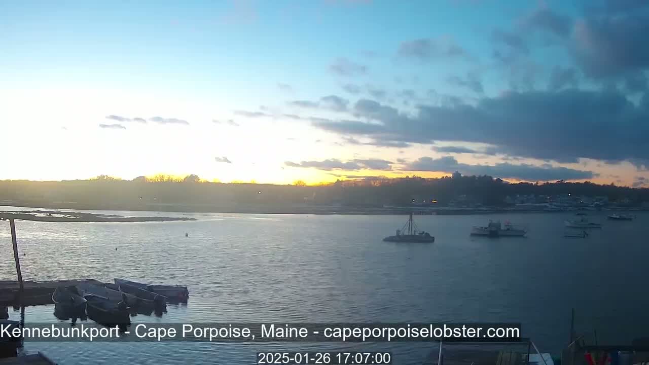 A calm body of water with several boats at a dock and a few others further out, under a partly cloudy sunset sky.