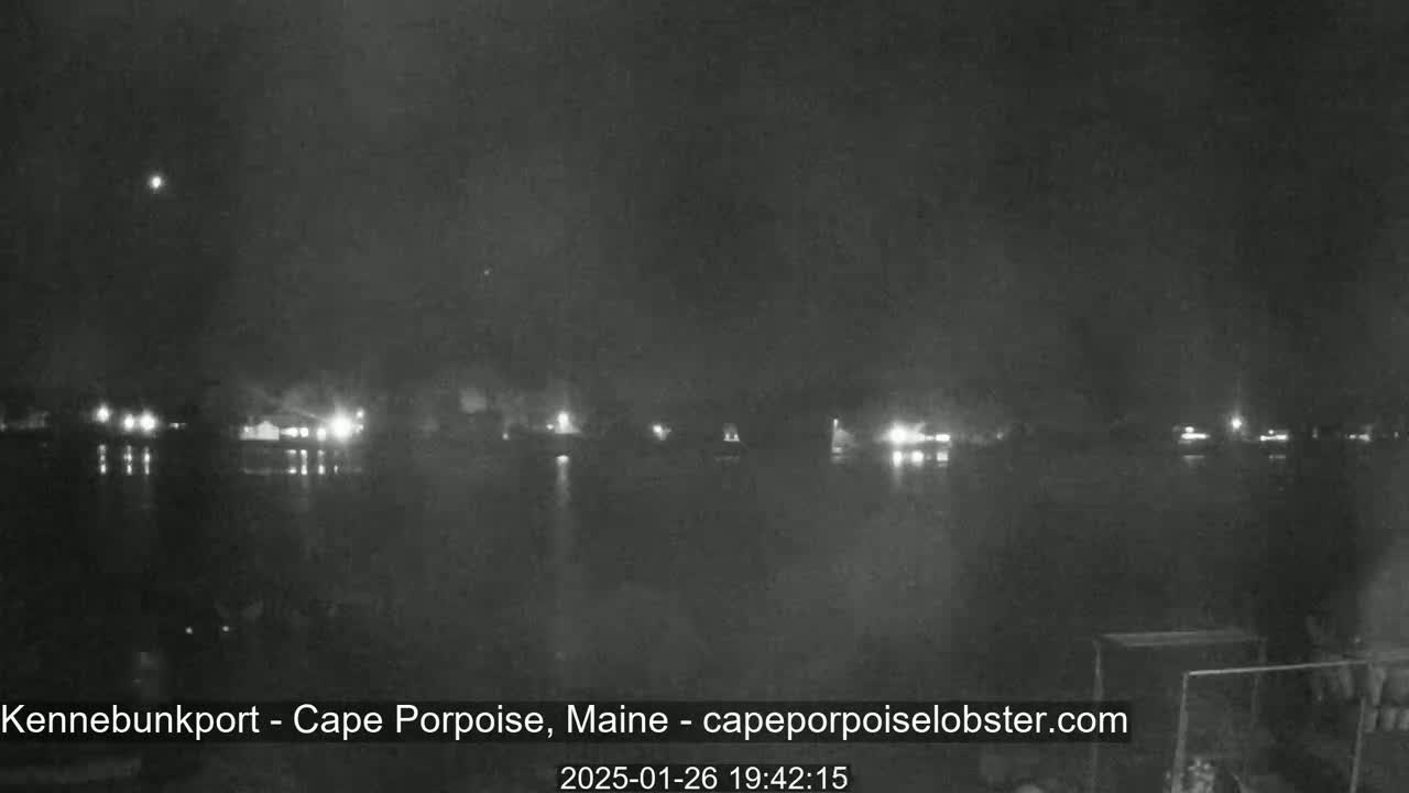A nighttime view shows a dark body of water reflecting the lights of a distant town under a clear, dark sky.