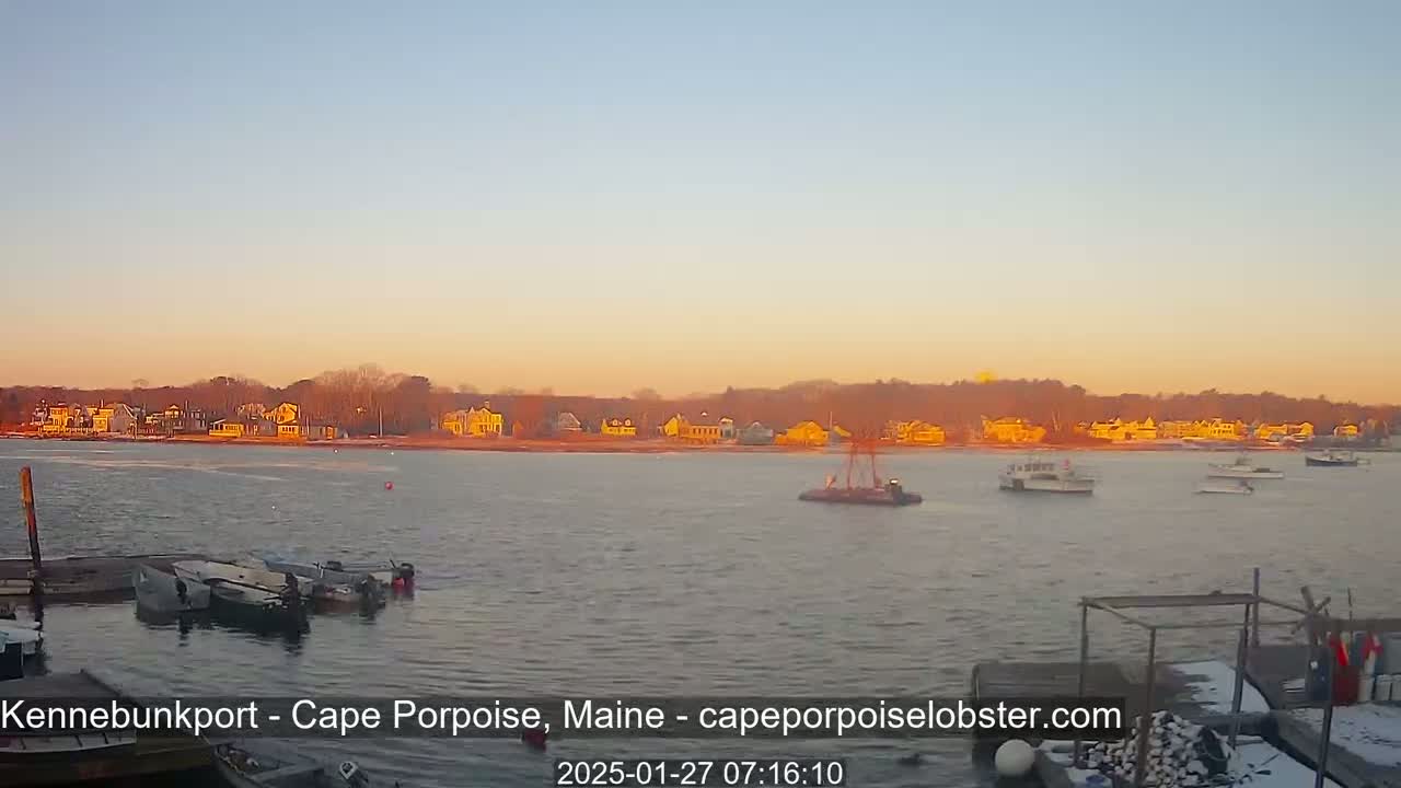 A calm body of water with several boats and a shoreline of houses bathed in the warm light of a sunrise or sunset.