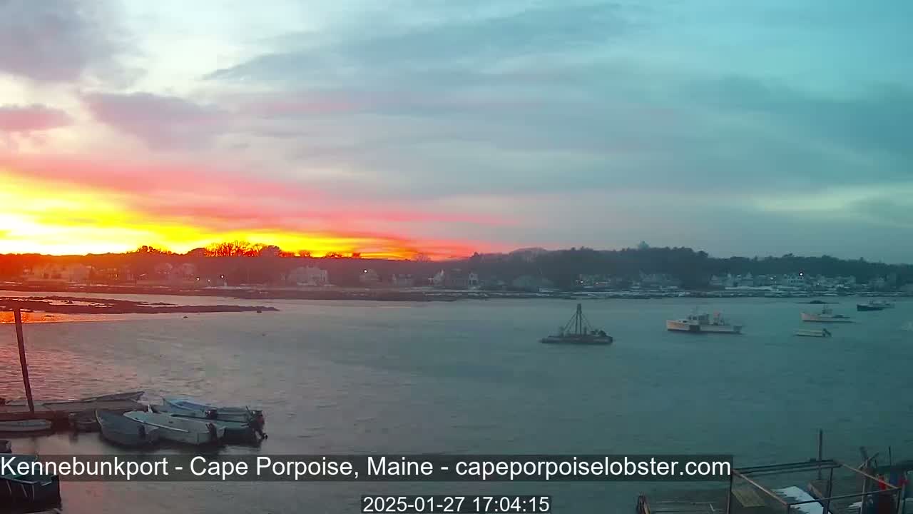 A vibrant sunset casts orange and pink hues across the sky above a calm body of water where several small boats are moored near a shoreline town.