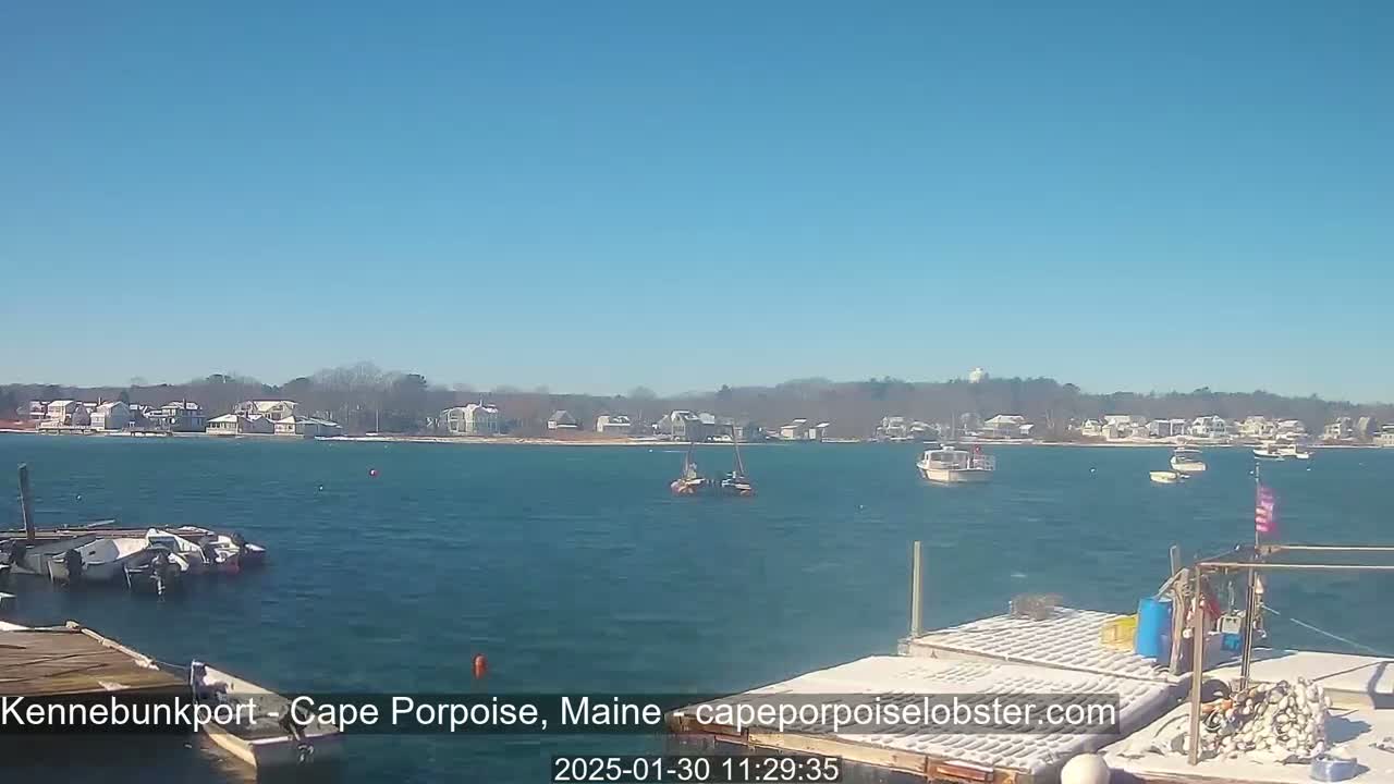 A calm, sunny day shows a harbor with several boats and a shoreline of houses with some snow visible on the ground and rooftops.
