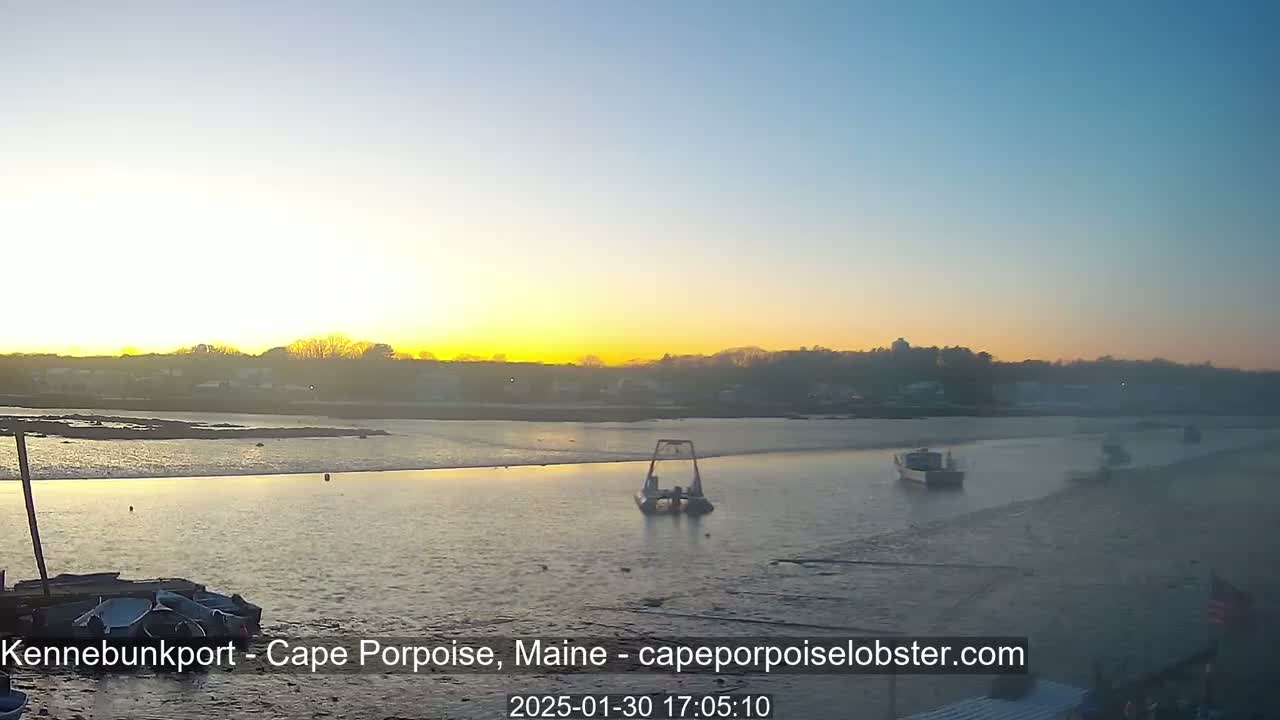 A calm body of water at sunset shows several boats, a metal structure in the water, and a shoreline with buildings and trees in the background.