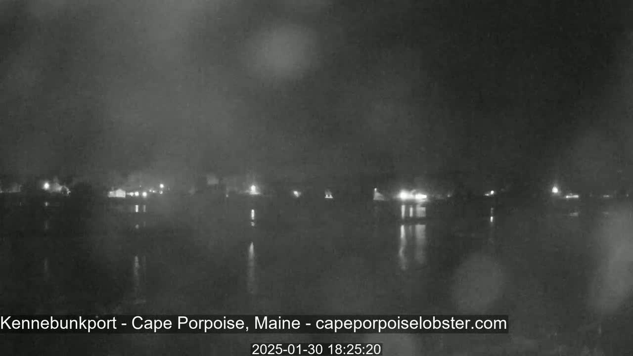 A nighttime, dimly lit view of a body of water reflecting the lights of a distant town under an overcast sky.