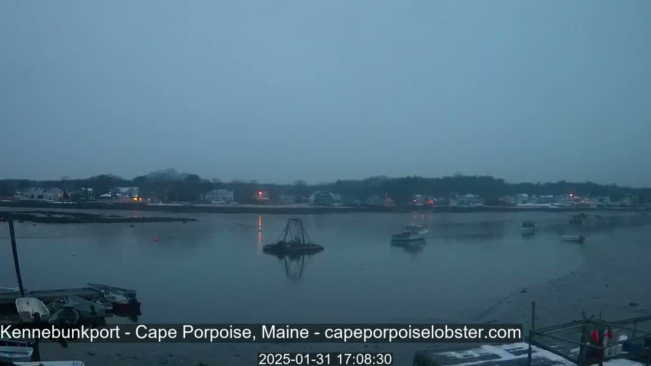 A calm body of water with several boats is seen in front of a snow-dusted town under an overcast sky.