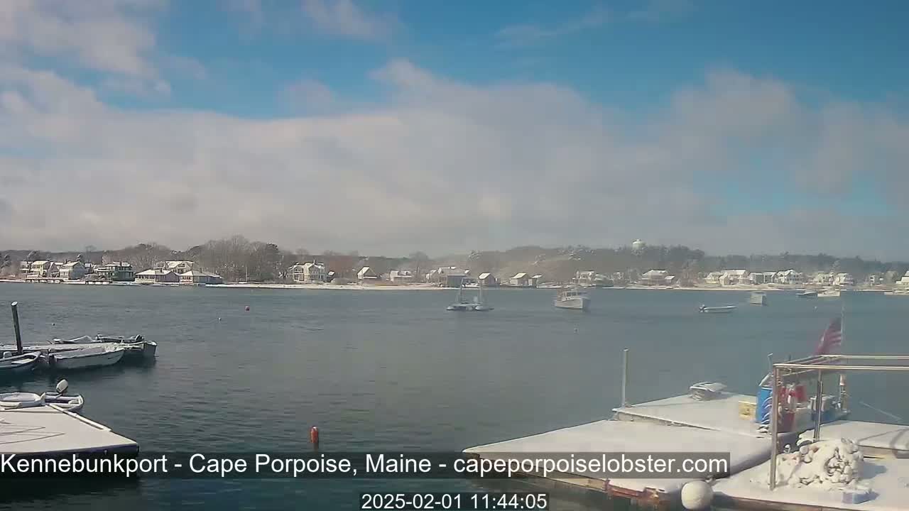 A partly cloudy, cold day shows a snow-covered waterfront town with boats in the harbor.